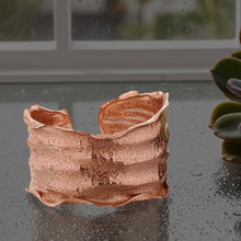 Load image into Gallery viewer, A copper-colored cuff bracelet with a textured, wavy design sits on a reflective surface, with a plant visible in the background.