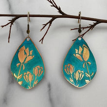 Load image into Gallery viewer, A pair of earrings with a floral design, featuring blue and gold colors, are hanging from a branch.