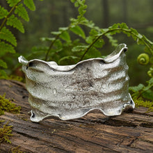 Load image into Gallery viewer, A silver cuff bracelet with a textured, wavy design rests on a wooden surface, surrounded by lush green foliage.