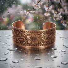 Load image into Gallery viewer, A copper-colored cuff bracelet with an intricate floral design rests on a wet surface, with water droplets scattered around it.