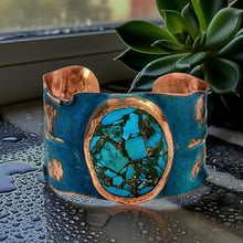 Load image into Gallery viewer, A turquoise and copper cuff bracelet with a large turquoise stone in the center, placed on a wet surface next to a green plant.