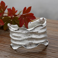 Load image into Gallery viewer, A silver cuff bracelet with a textured, wavy design sits on a wooden surface, with red autumn leaves in the background.
