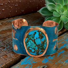 Load image into Gallery viewer, A blue and copper cuff bracelet with a large turquoise stone in the center, placed on a wooden surface with a green succulent plant in the background.
