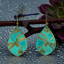 Load image into Gallery viewer, A pair of turquoise earrings with gold designs, including a dragonfly, are displayed on a branch with a purple flower.
