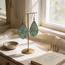 Load image into Gallery viewer, A pair of turquoise earrings with gold leaf designs, displayed on a gold stand, with a small bowl containing rings and a plant leaf nearby.
