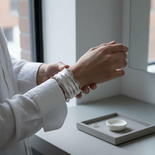 Load image into Gallery viewer, A person is adjusting a silver bracelet on their wrist, with a small white bowl and a mirror in the background.

