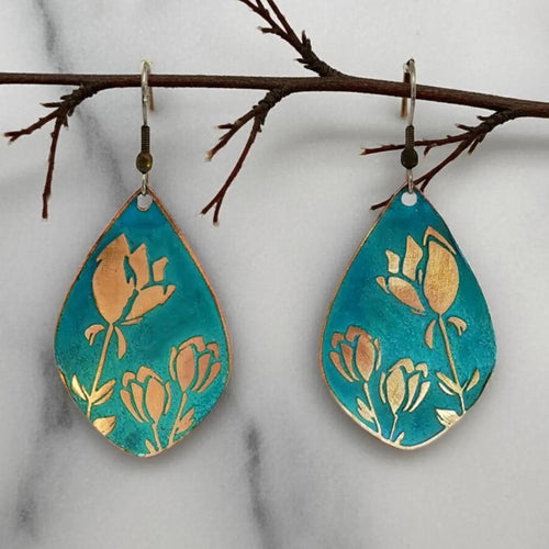 A pair of earrings with a floral design, featuring blue and gold colors, are hanging from a branch.