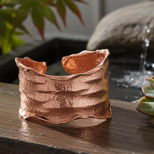 Load image into Gallery viewer, A copper-colored cuff bracelet is displayed on a wooden surface, with a blurred background featuring plants and a rock.
