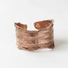 Load image into Gallery viewer, A copper-colored cuff bracelet with a textured, wavy design sits on a plain white background.
