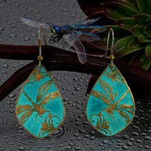 Load image into Gallery viewer, A pair of turquoise earrings with gold designs, including a dragonfly, are displayed on a branch with a green plant in the background.
