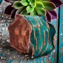 Load image into Gallery viewer, a close-up of a copper-colored, textured bracelet with a greenish-blue hue, placed on a wooden surface with water droplets around it. In the background, there is a green plant with purple petals, creating a visually appealing contrast.
