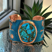 Load image into Gallery viewer, A blue and copper cuff bracelet with a large turquoise stone in the center, placed on a glass surface with water droplets on it.
