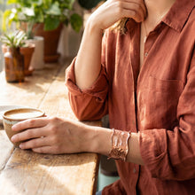 Load image into Gallery viewer, A person wearing a red shirt is sitting at a wooden table, holding a cup and resting their hand on their chin.
