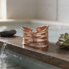 Load image into Gallery viewer, a copper bracelet resting on a wooden ledge, with a small succulent plant and a rock nearby, all situated in a tranquil setting with a body of water.
